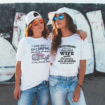 Two women wearing ball caps and humorous white graphic tees standing against a grungy background, embodying a fun and laid-back style.