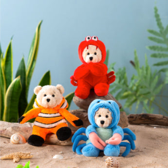 Three teddy bears dressed in colorful animal costumes on a sandy surface with plants in the background.