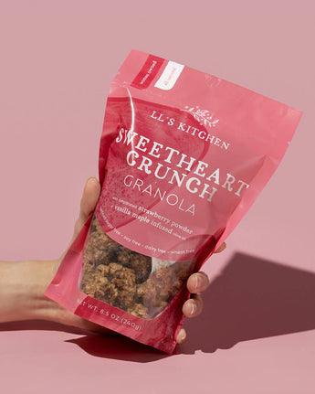 Hand holding a pink package of 'Sweetheart Crunch Granola' by LL's Kitchen against a pink background
