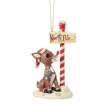 Jim Shore Rudolph by North Pole Sign ornament featuring Rudolph wearing a Santa hat beside a candy cane striped North Pole sign.