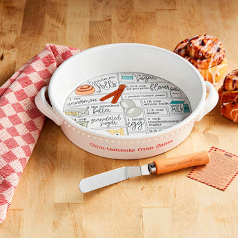 Cinnamon Roll Recipe Baker Set featuring a ceramic dish with recipe printed inside, holiday spatula, and recipe card, styled with cinnamon rolls and red dish towel.