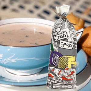 A bowl of She Crab Soup with a packaged soup mix in the foreground, set on a table with silverware and a napkin.