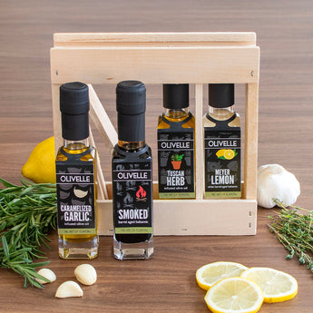 Set of Olivelle olive oil bottles and balsamic vinegars in a wooden gift box with lemons and herbs on a wooden surface.