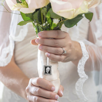 A close-up of a woman's hand holding a pink bouquet of flowers with a wedding bouquet memorial charm that includes a picture frame, primarily silver in color.