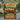 A rectangular metal-style sign with rounded corners stands outdoors on a gravel path near campers and trees. The sign features a vintage-style illustration of a camper trailer, a folding chair, a campfire, mountains, and large text reading “WELCOME TO OUR CAMPER,” “WE HAVE BEER AND A HOME ON WHEELS,” and “WHAT COULD POSSIBLY GO WRONG?”.