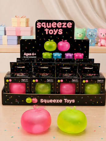 A retail display of squeeze toys in black boxes labeled 'squeeze toys' and 'Ages 6+', with two large translucent apple-shaped toys in front: one pink and one lime green. Background shows a soft beige curtain and more colorful plush bears and gift boxes on a shelf.