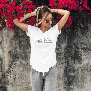 A person standing by a wall with red flowers, wearing a white crew neck t-shirt with 'South Dakota' text and a heart symbol.