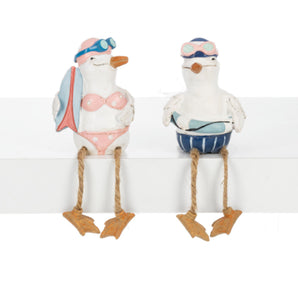 Two ceramic figurines of seagulls, one wearing a bikini and the other in swim trunks with a floatie, displayed on a shelf.