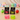 Two boxed apple-shaped squeeze toys in front of their black retail packaging, with two loose apple squeezers in pink and green on the table. A wooden shelf in the background holds colorful gift boxes and small bear figurines.