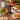 A bottle of Olivelle Sun-dried tomato barrel aged balsamic stands on a wooden table surrounded by prepared food, including a pizza, pasta, salad, bread, and bowls of oil and sunflower oil, with a basket of fresh vegetables in the background.