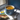 Blue mug filled with soup on a windowsill next to a spoon and packets of instant noodles.