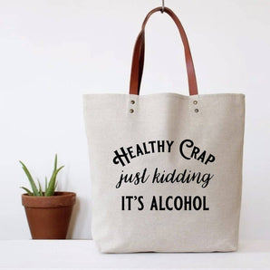 A beige cotton tote bag with brown faux leather handles and the text 'HEALTHY CRAP JUST KIDDING IT'S ALCOHOL' printed on the front.
